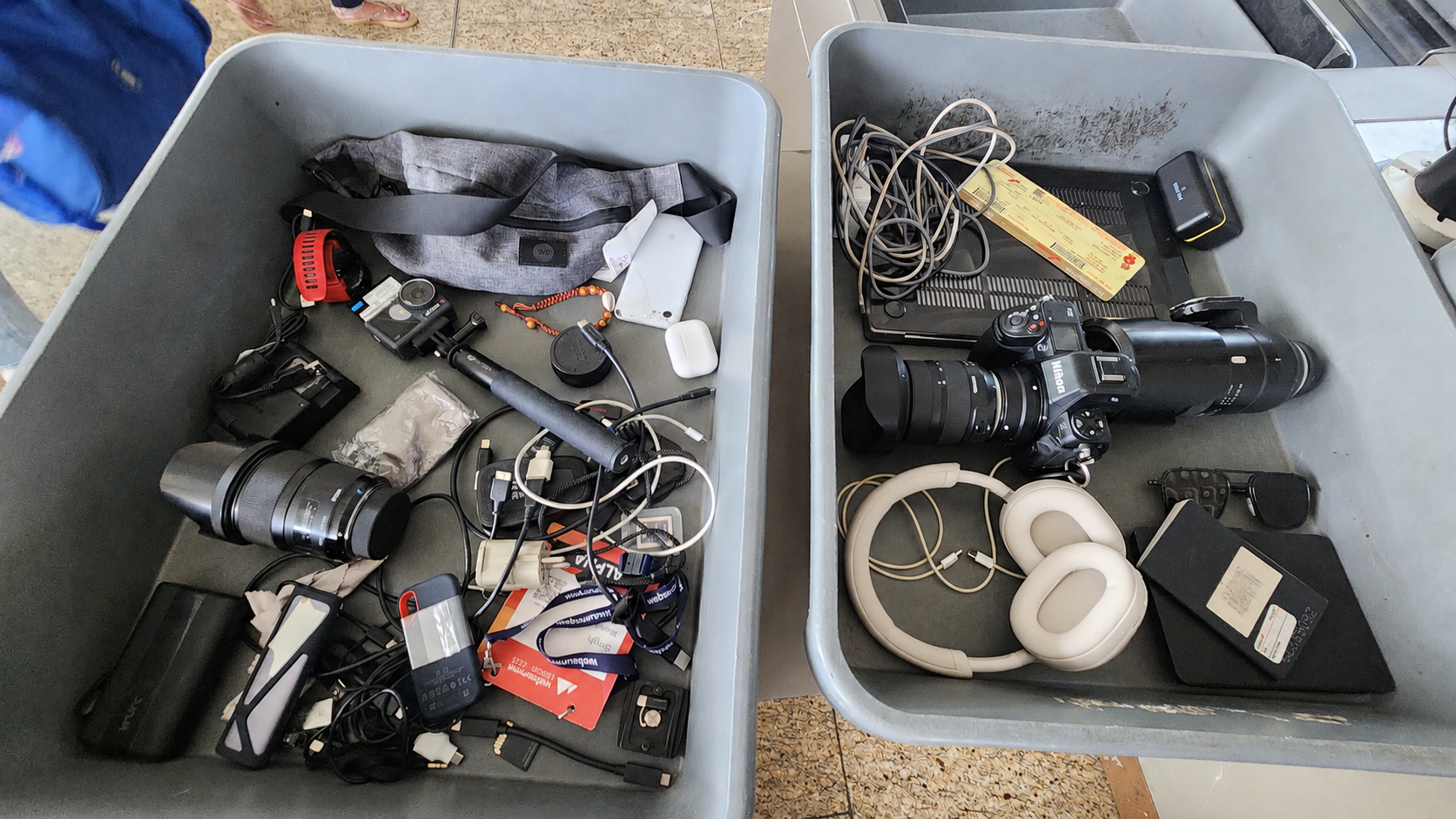 Two airport-security trays overflowing with a Nikon DSLR, an action cam, headphones, a sports watch, SSDs, batteries, and a tangle of cables